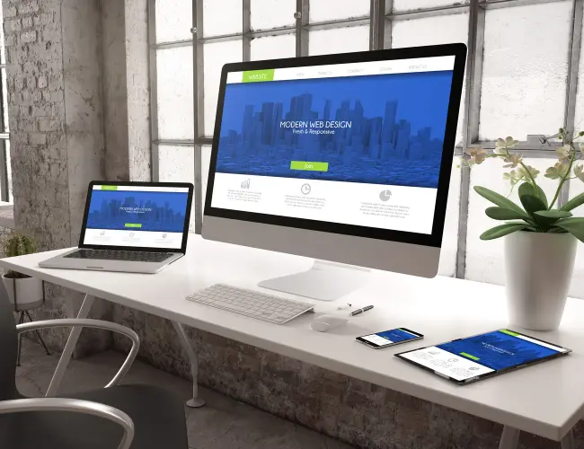 A laptop, desktop computer, phone, and tablet all spread out on a work desk, displaying the same website, as an example of responsive web design