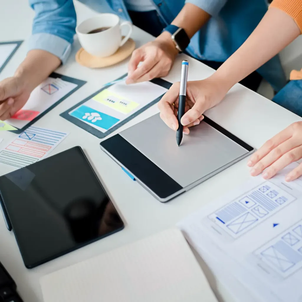 A pair of web designers creating graphics with a drawing tablet, with planning papers on website design scattered around the table they're working on