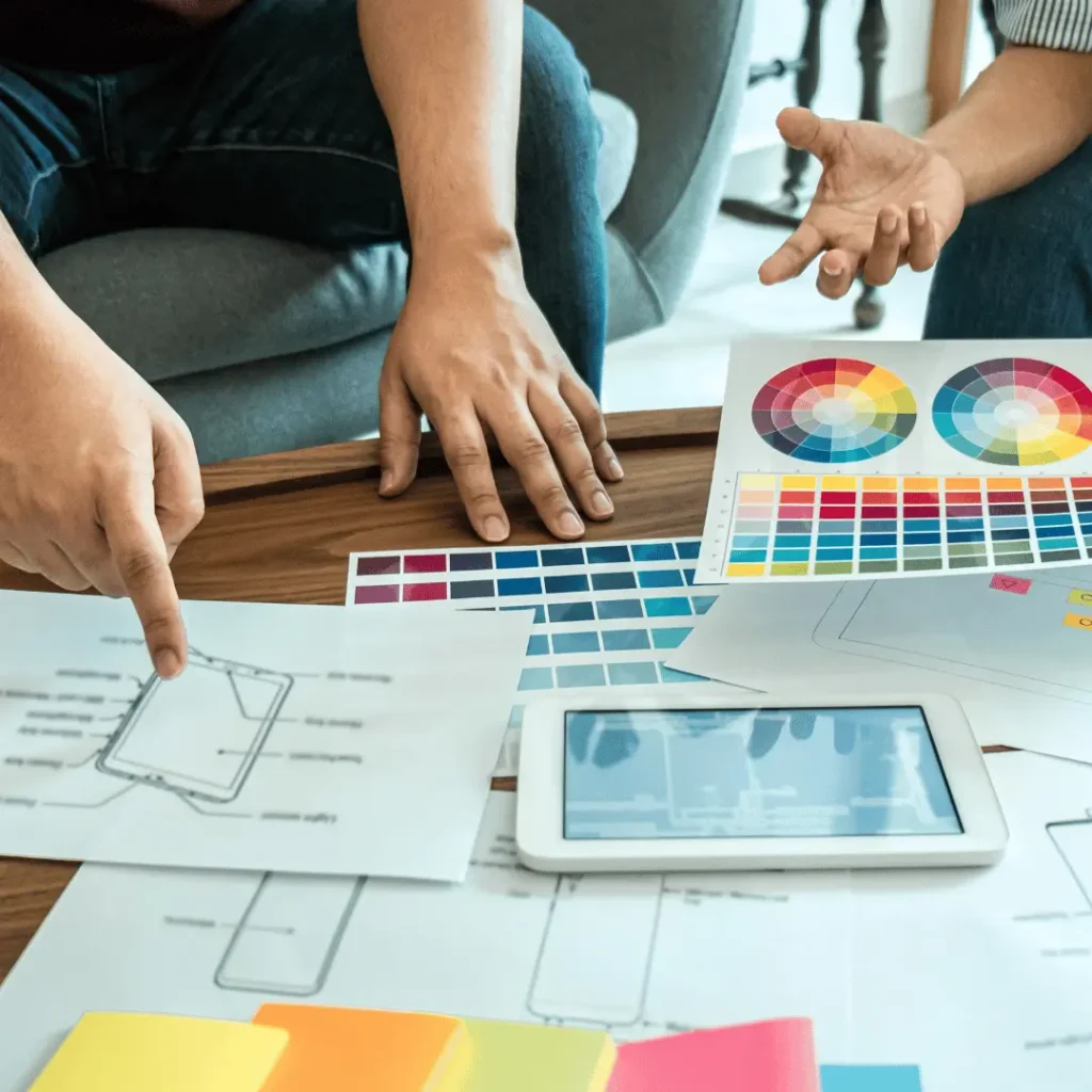 A pair of people working together on responsive web design, with design drafts and colour schemes scattered over the desk they're working on