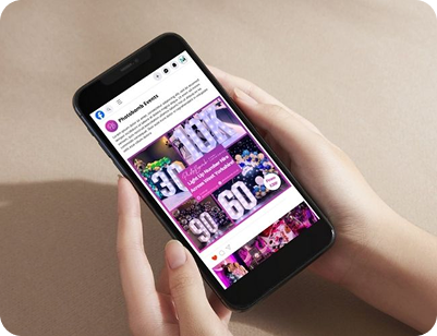 A pair of hands holding a smartphone showing a Facebook business page for Photobomb Events, showcasing a collage of light-up number rentals for various celebrations.