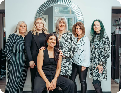 A group of six diverse female stylists posing together in a bright, modern salon in front of a circular mirror.