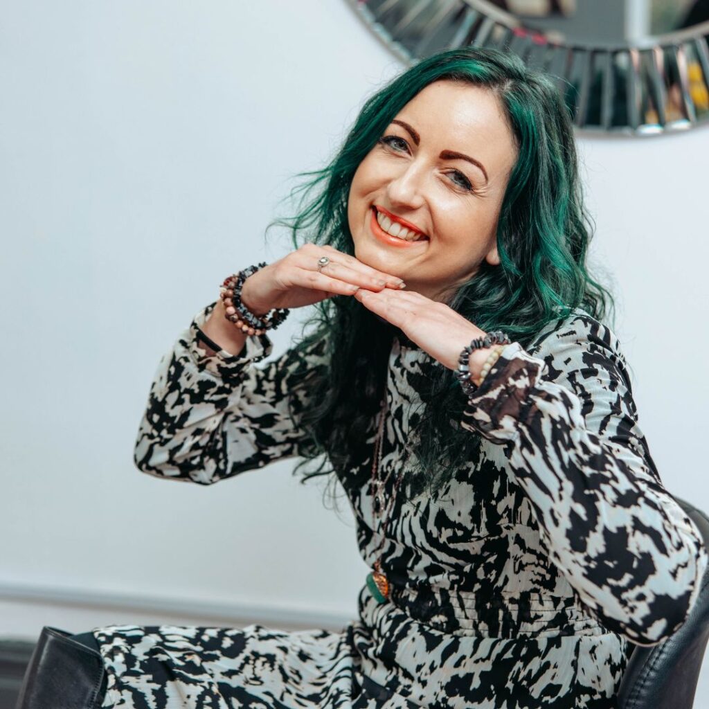 A smiling woman with green hair resting her chin on her hands while seated indoors.