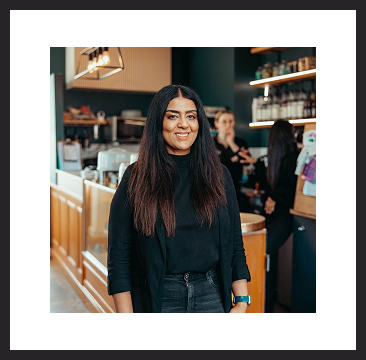 A woman standing inside a café, smiling confidently, with the coffee counter and staff working in the background.