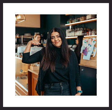 A woman smiling inside a café, pointing to herself with the counter and shelves behind her.