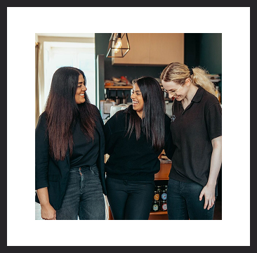 Three women standing together inside a café, smiling and laughing with their arms around each other.