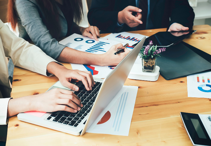 A close-up, high-angle view of a team collaborating at a wooden desk, featuring hands typing on a laptop and pointing at printed financial charts and pie graphs.