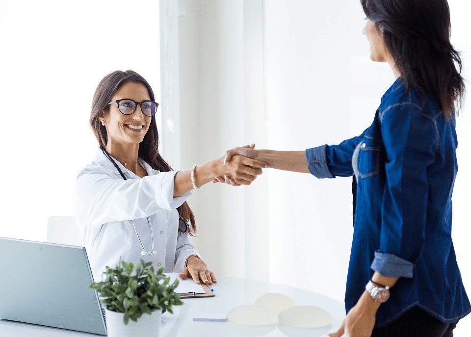 A smiling female doctor in a white coat and glasses shaking hands with a female patient across a white desk in a bright medical office.