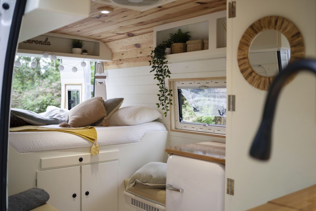 Cozy camper van interior with a bed, wooden ceiling, plants, and compact kitchen area.