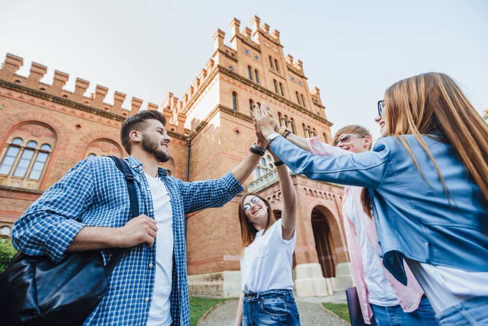 Group of university students giving a high five in front of a historic campus building.