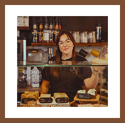 Barista standing behind a café counter with cakes on display.