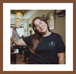 A smiling café worker wearing glasses and a Café 29 t-shirt playfully flexes her arm inside the café.