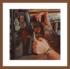 A tattooed barista steams milk using an espresso machine.