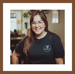 A woman wearing glasses and a black Café 29 T-shirt smiles at the camera indoors.