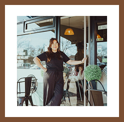 A woman smiling while standing in the doorway of a café, with outdoor seating and greenery beside her.