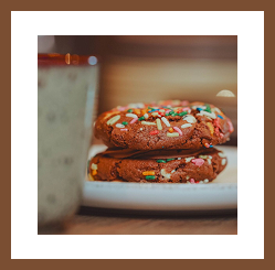 Two chocolate cookies with colourful sprinkles stacked on a plate, with a coffee cup blurred in the foreground.