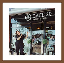 A woman stands outside Café 29 on Anders Square, smiling and gesturing toward the café entrance.