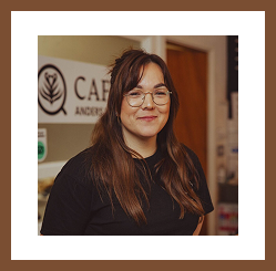 A smiling woman with glasses and long hair stands indoors, with café signage visible behind her.