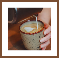 Milk is being poured into a ceramic cup of coffee, creating latte art on the surface.