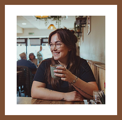A smiling woman wearing glasses sits inside a café, holding a cup of coffee and looking to the side.