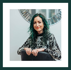Portrait of a woman with teal hair seated indoors, smiling confidently at the camera