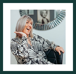 Portrait of a smiling grey-haired woman seated indoors, playfully gesturing with her hand