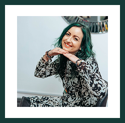 A smiling woman with green hair resting her chin on her hands while seated indoors.