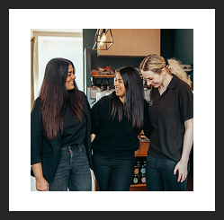 Three women standing together, smiling and talking in a café or workspace.