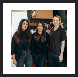Three women standing together and smiling in a café or workspace.